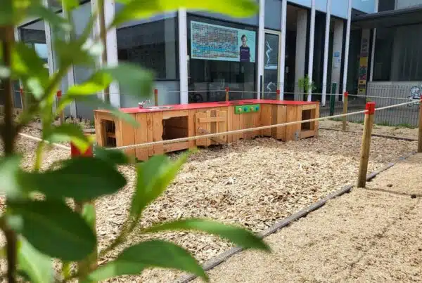 Houten kinderkeuken op een schoolplein, omgeven door houtsnippers en jonge bomen. Voorbeeld van duurzame speel- en leeromgevingen.