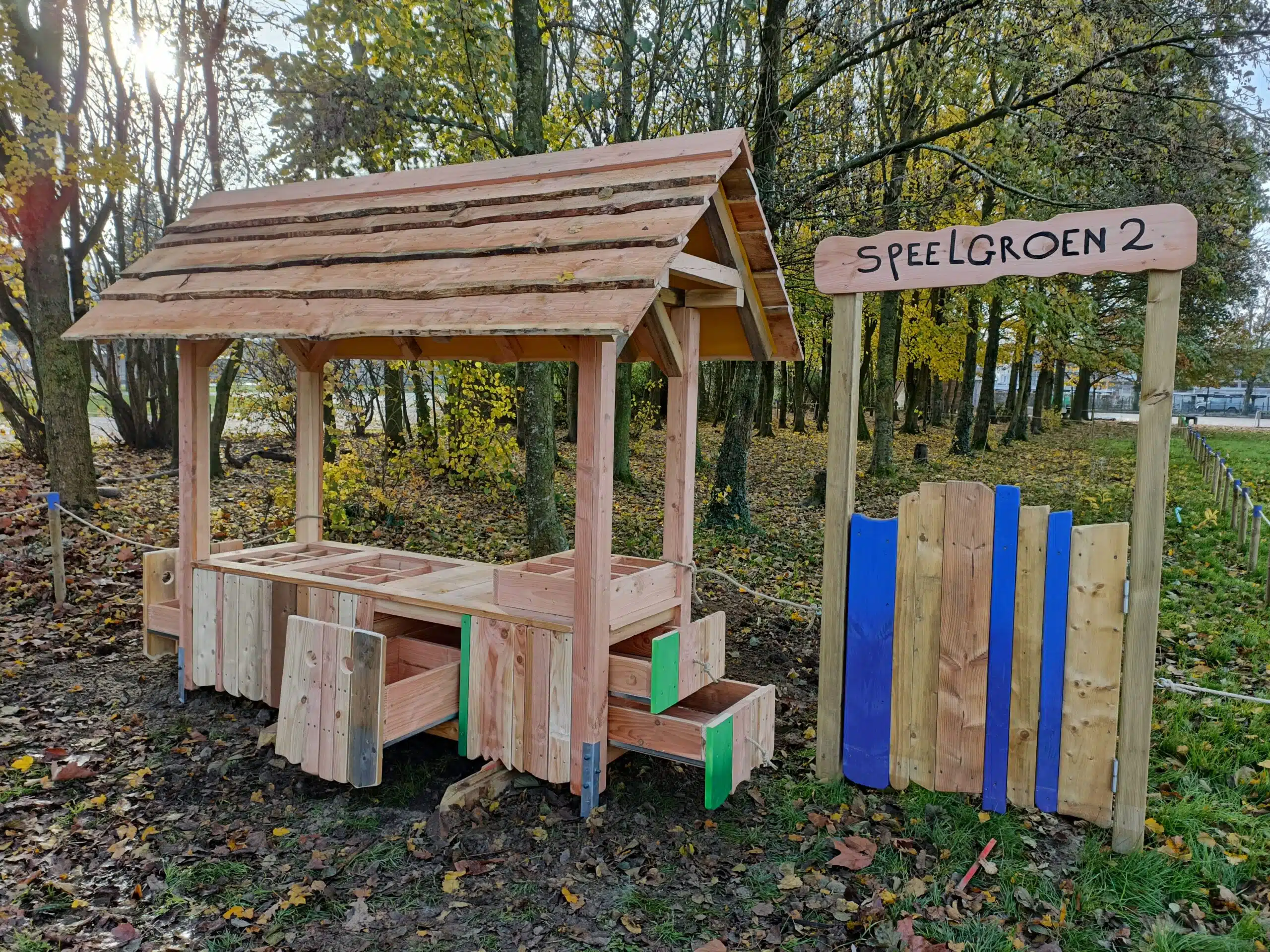 Houten superette met dak en open tafeloppervlak, omgeven door bomen en herfstbladeren, met een bord "Speelgroen 2".