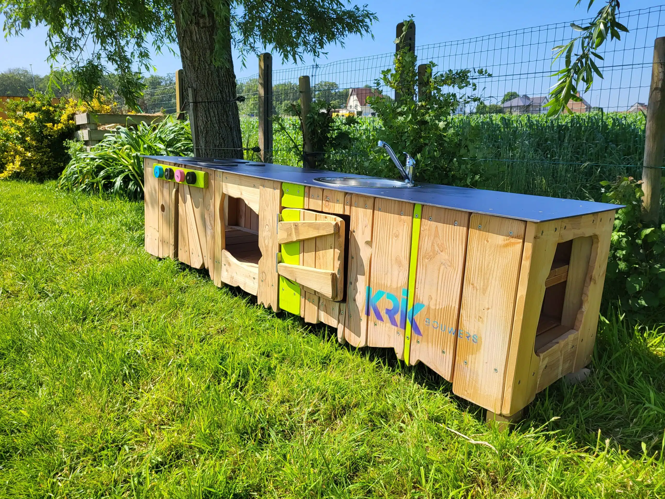 Houten kinderkeuken met gootsteen en kleurrijke knoppen, geplaatst in een groene tuinomgeving.